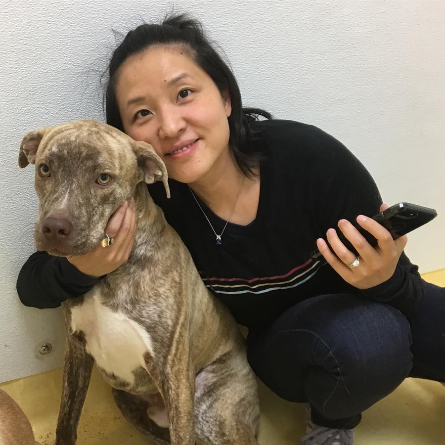 Jen crouched against a white wall, her arm around Tiger, a young brindle dog who leans into her.